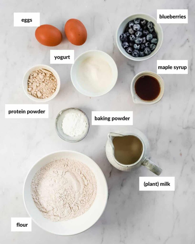 all ingredients needed for protein pancakes without banana: eggs, maple syrup, plant milk, yoghurt, flour, protein powder, baking powder, salt, blueberries.