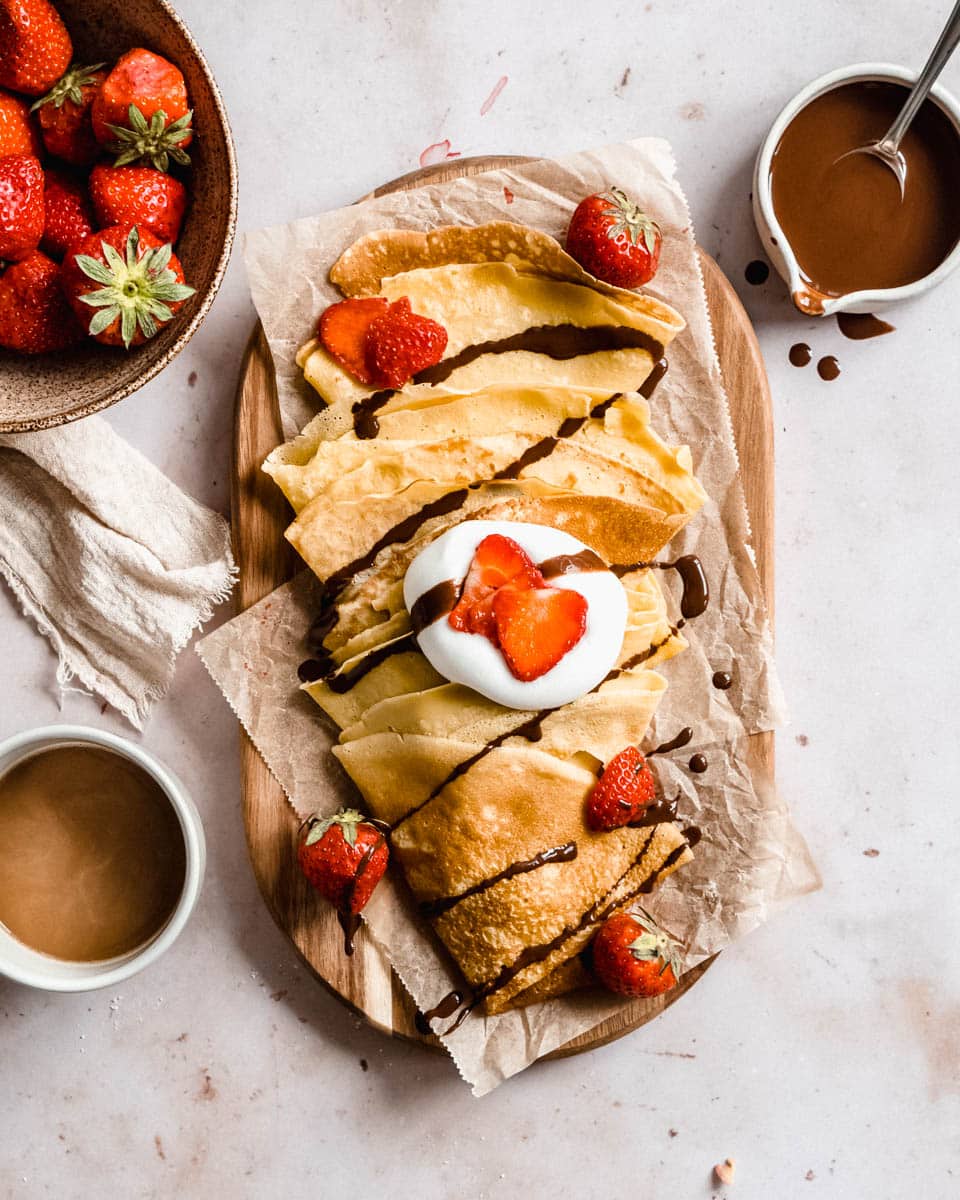 stack of almond milk crepes on a wooden board topped with fresh strawberries and vegan whipped cream, next to it some melted chocolate, fresh strawberries and coffee.