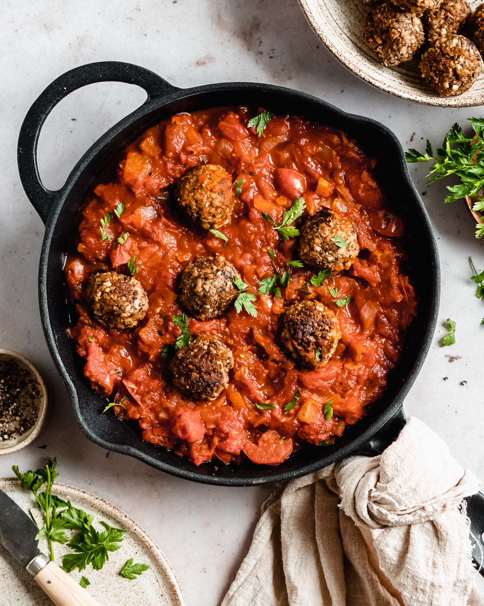 6 veggie meatballs in marinara sauce in a skillet, next to it some fresh herbs and black pepper.