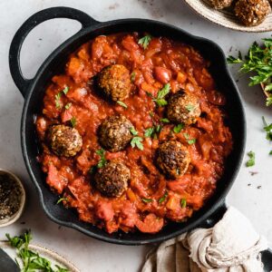 6 veggie meatballs in marinara sauce in a skillet, next to it some fresh herbs and black pepper