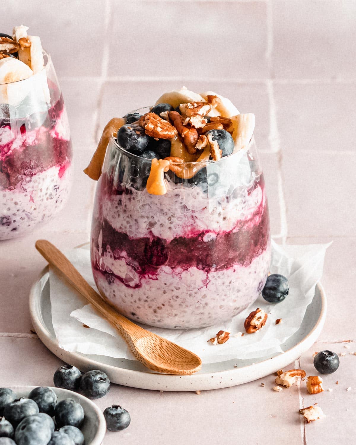 overnight oats with frozen blueberries in a glass, in the middle one layer of mashed blueberries and topped with fresh blueberries, banana, nuts and caramel sauce.