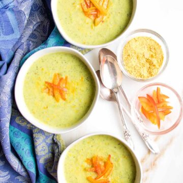 Three white bowls, filled with light green creamy zucchini soup and sprinkled with nutritional yeast and strips of red pepper on a white background with a blue tea towel.