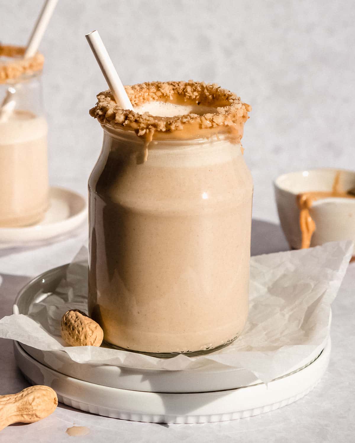 2 glasses of peanut butter smoothie, one up close, with a straw and some peanuts next to it.