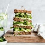 big guacamole sandwich filled with cucumber, lettuce and micro greens on a wooden plate.