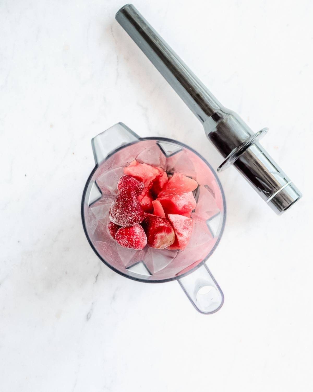 maple syrup, watermelon chunks ,frozen strawberries in a blender with a tamper next to it.