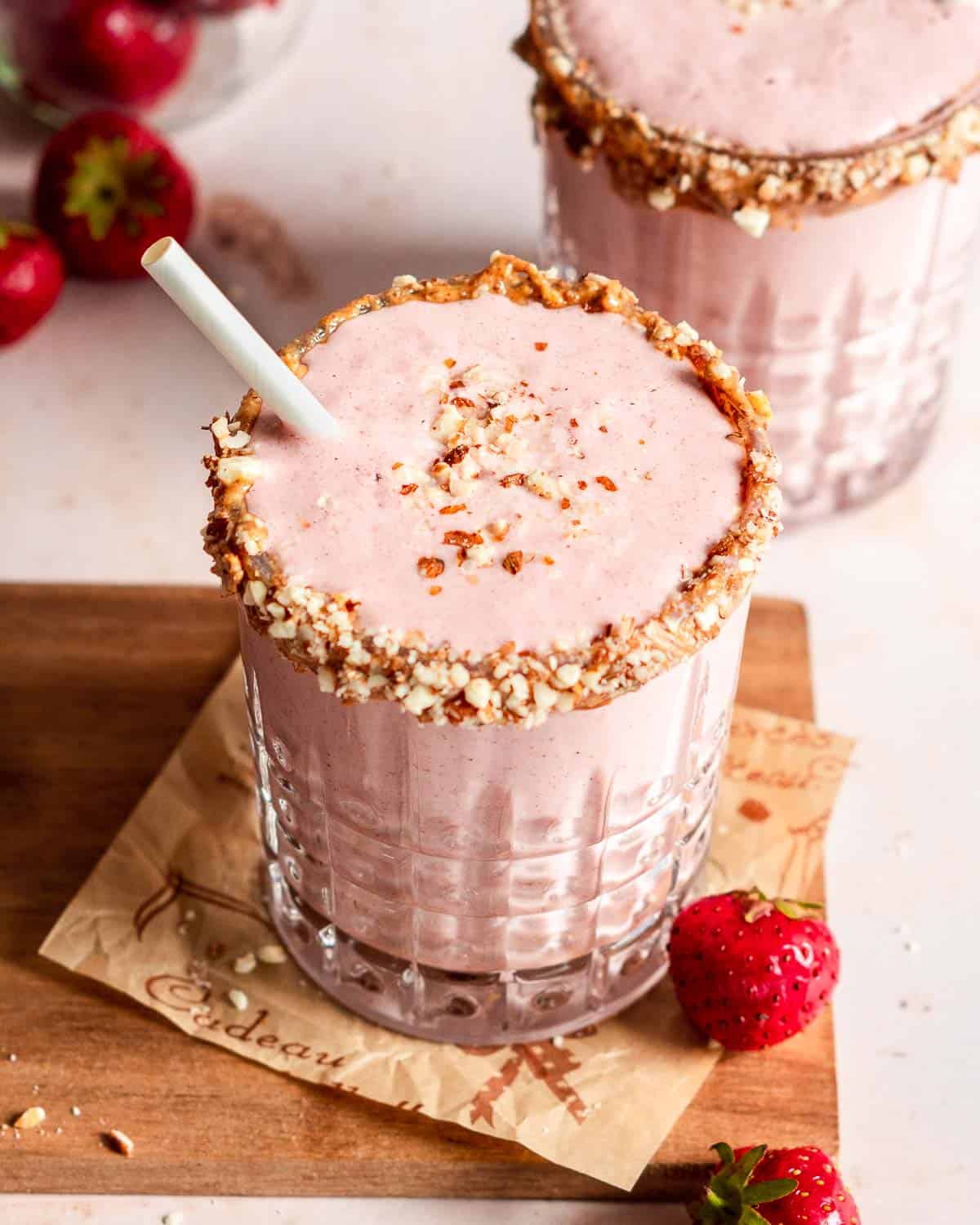 2 serving glasses of strawberry smoothie with a straw on a wooden palte, with some fresh strawberries next to it.