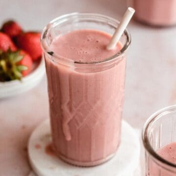 1 serving glass of strawberry smoothie on a marble plate, next to it a second smoothie and some fres strawberries.