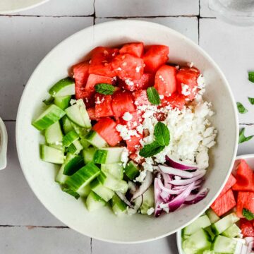 close up of big bowl of watermelon salad, next to it some dressing, a glass of water.