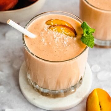 serving glass of frozen peach smoothie topped with fresh peach slices, mint leaves and a straw.
