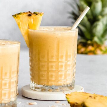 2 glasses of frozen pineapple smoothie with a straw. Pinapple head in the back, some fresh pineapple at the front.
