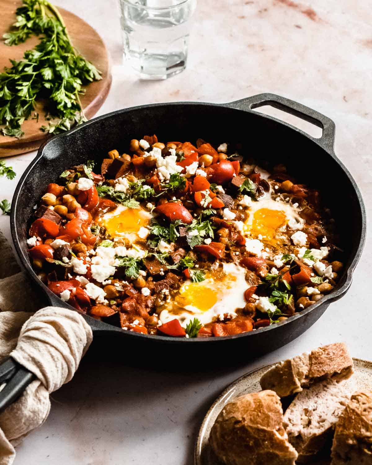 shakshuka in a big cast iron pan with three eggs, next to it some bread and fresh parsley and a glass of water.