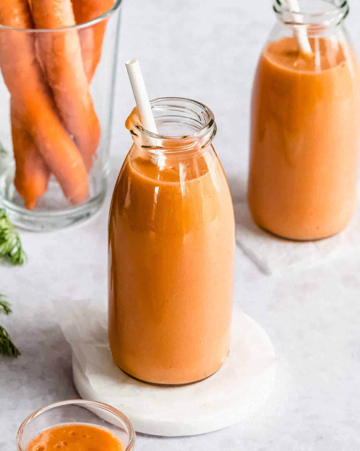 2 glasses of carrot smoothie with a straw, next to it some carrots.