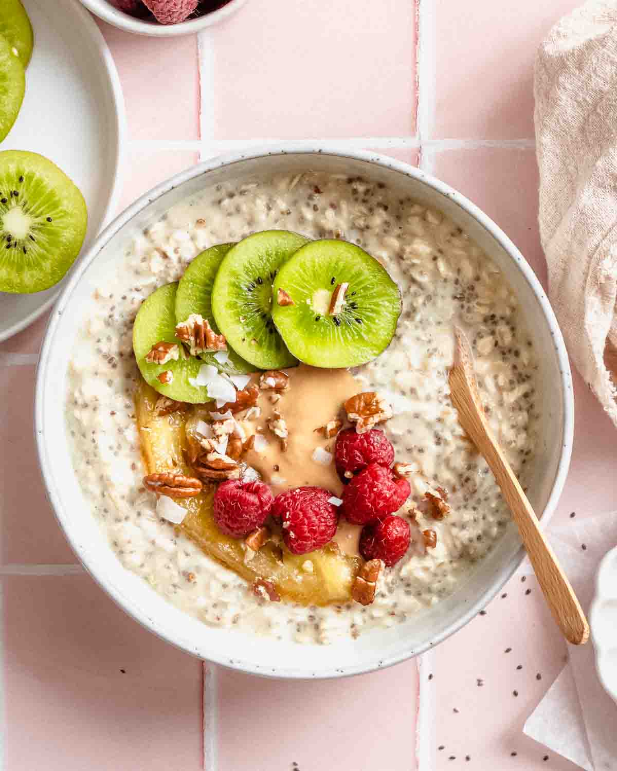 overnight oats in a bowl topped with kiwi, raspberries, caramelized banana and peanut butter., homemade granola and coconut flakes.
