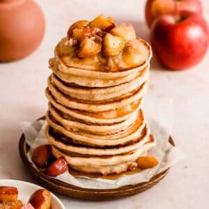 stack of vegan applesauce pancakes on a wooden plate topped with caramelized apples, nuts and applesauce.