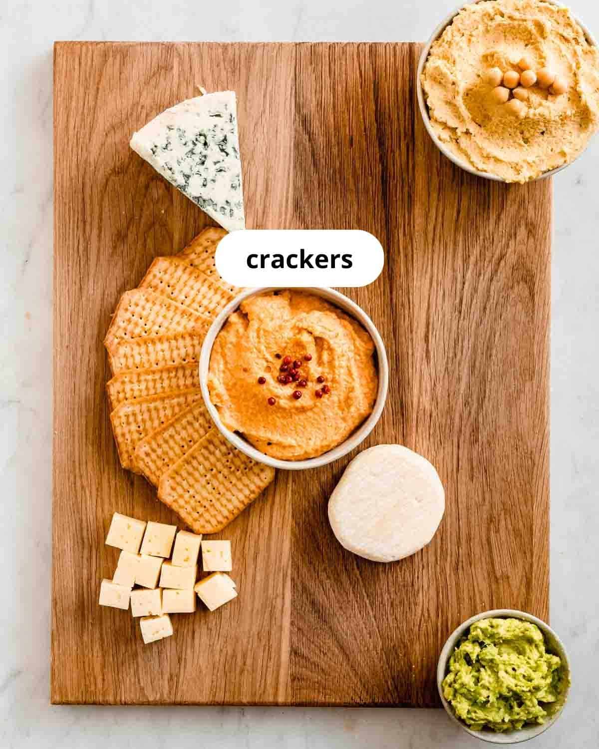 cutting board with hummus, a spicy spread, guacamole, and 3 types of cheese and crackers on it.