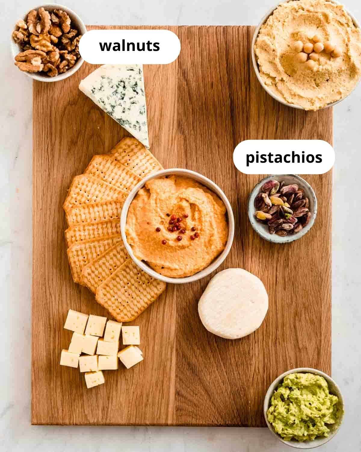 cutting board with hummus, a spicy spread, guacamole, and 3 types of cheese, crackers, walnuts and pistachios on it.