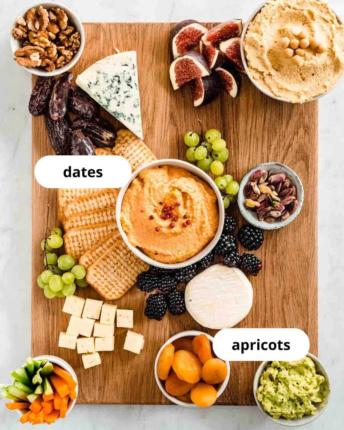 cutting board with hummus, a spicy spread, guacamole, and 3 types of cheese, crackers, walnuts, pistachios, carrot and celery sticks, fresh figs, grapes, berries, dates and apricots on it.