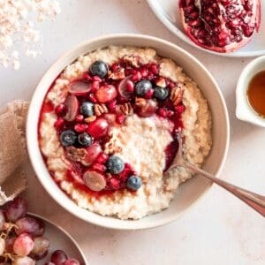 millet porridge in a bowl topped with fresh fruit. next to it fresh pomegranate, maple syrup and grapes.