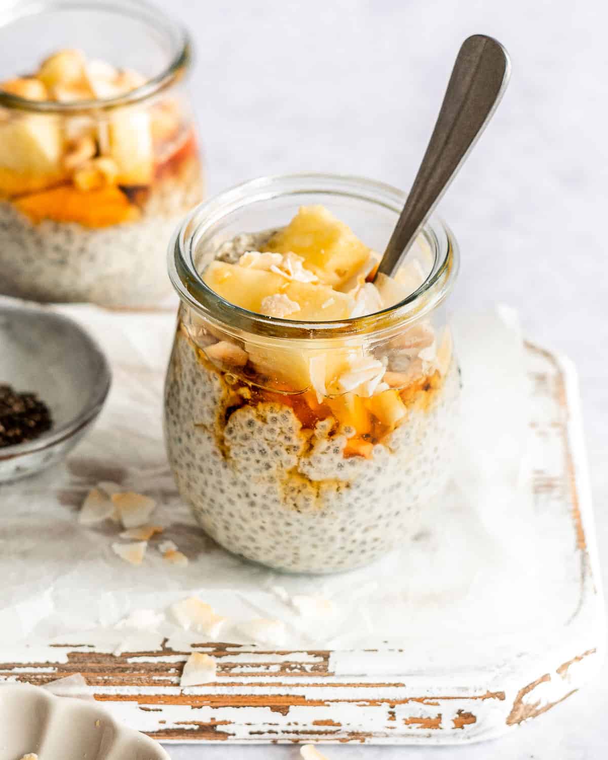 coconut milk chia seeds in a small jar with a spoon in it topped with fresh pineapple, nuts, coconut flakes and maple syrup.