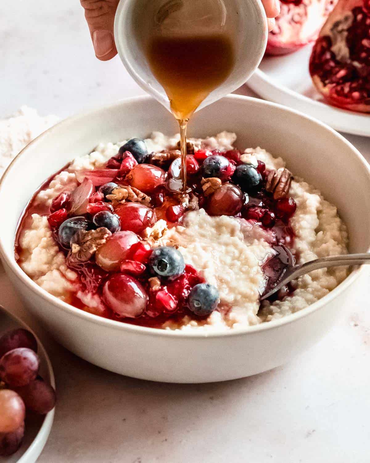 millet porridge in a bowl topped with blueberries, grapes, pomegranate seeds and pecans.. maple syrup being drizzled on.