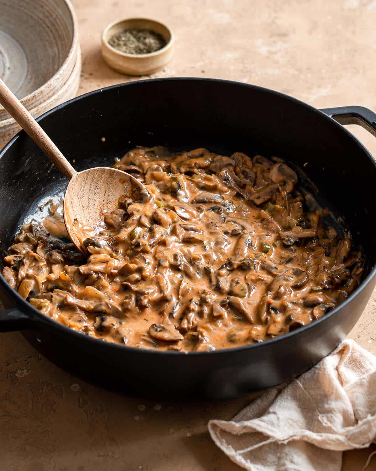 1 cast iron skillet filled with ragout of mushrooms and a wooden spoon next to it a linen napkin and 2 plates.