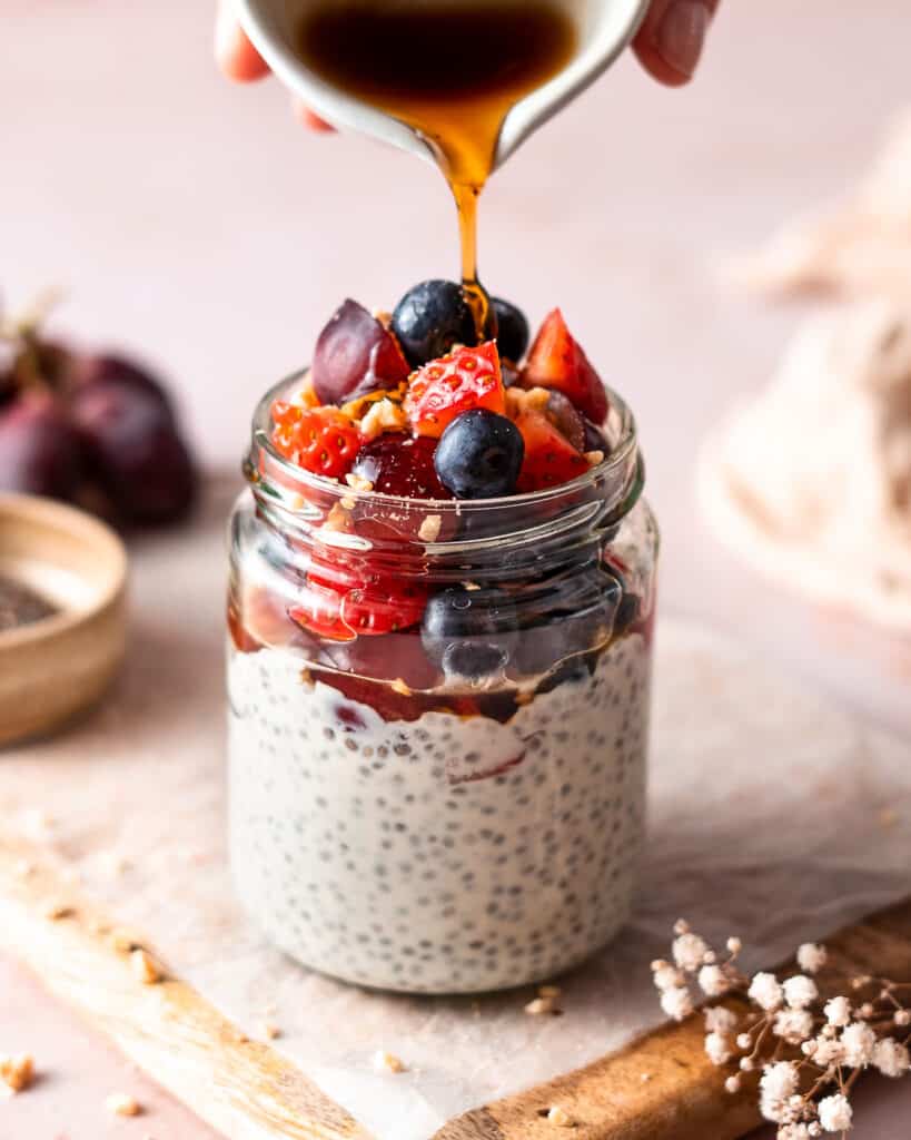 yogurt chia pudding in a glass jar on a cutting board, topped with fresh fruit and maple syrup.