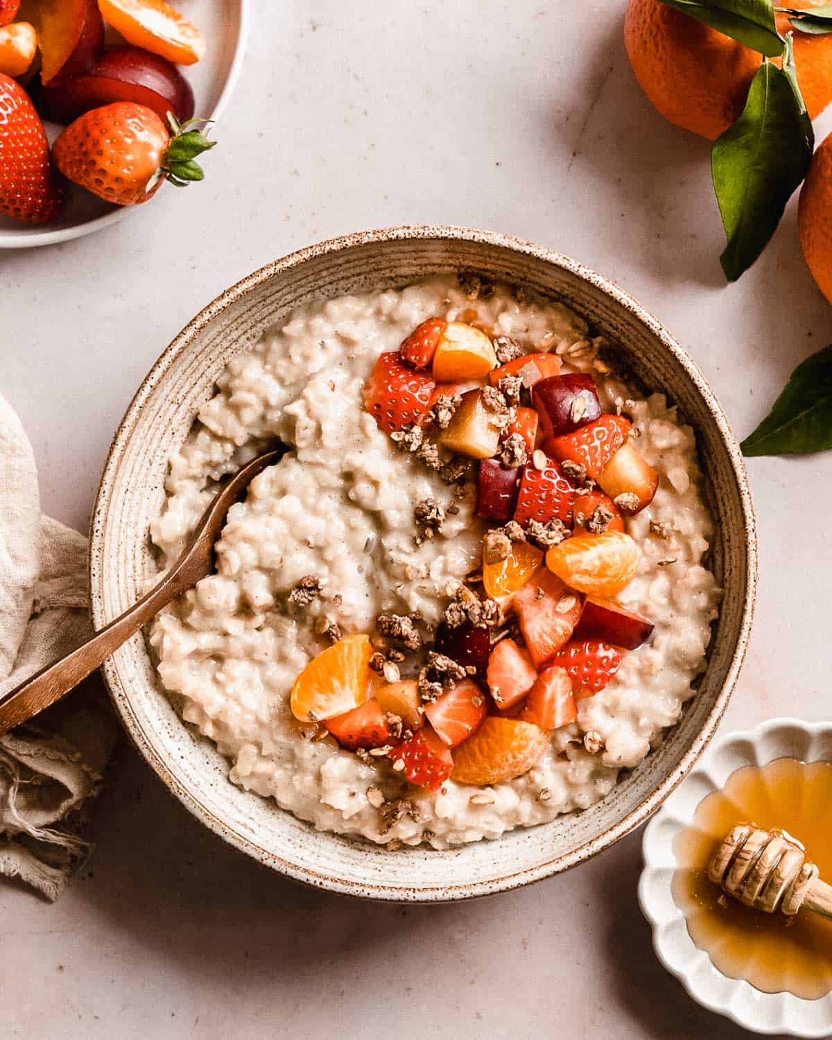 honey oatmeal in a bowl topped with fresh fruit and granola. next to it some fresh fruit and honey.