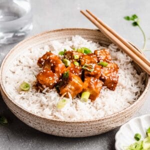 sticky tofu topped with sesame seeds on a bowl of white rice with chopsticks.