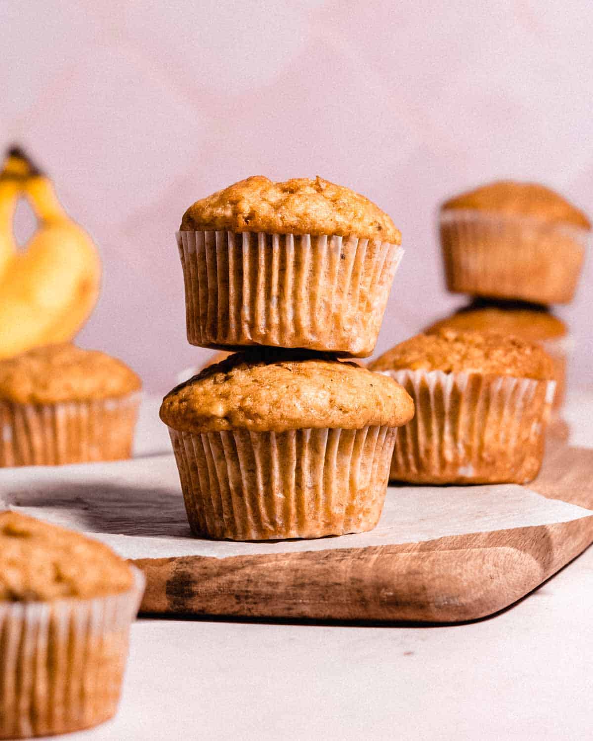 stack of 2 oil banana muffins on a wooden cutting board, in the background more muffins and 2 bananas.