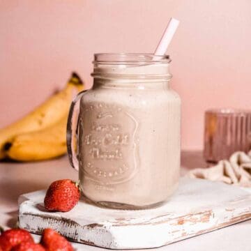 high calorie smoothie in a glass jar with a straw. behind some resh bananas and in front of it strawberries.