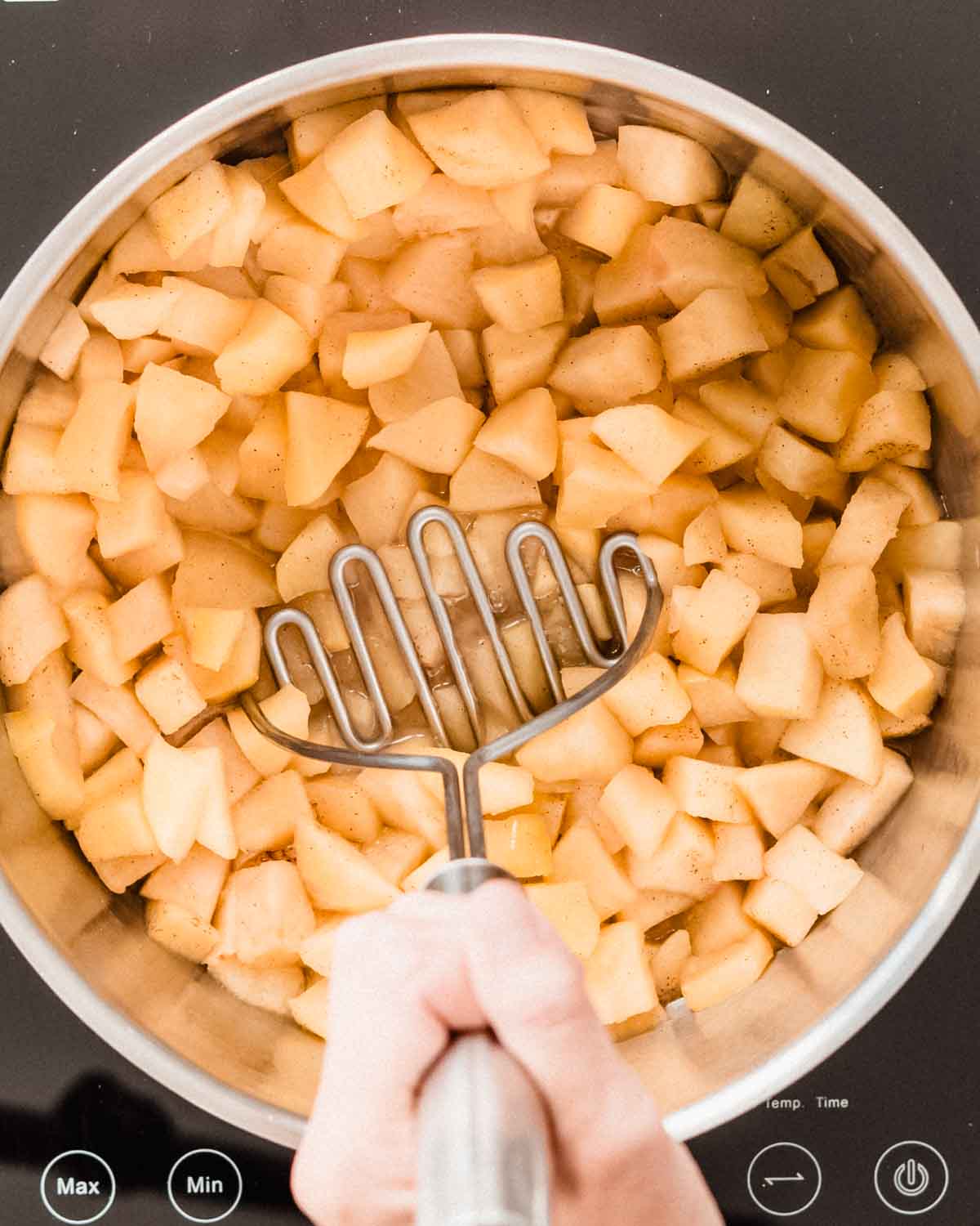 Cooked potato chunks in a saucepan being mashed with a potato masher.