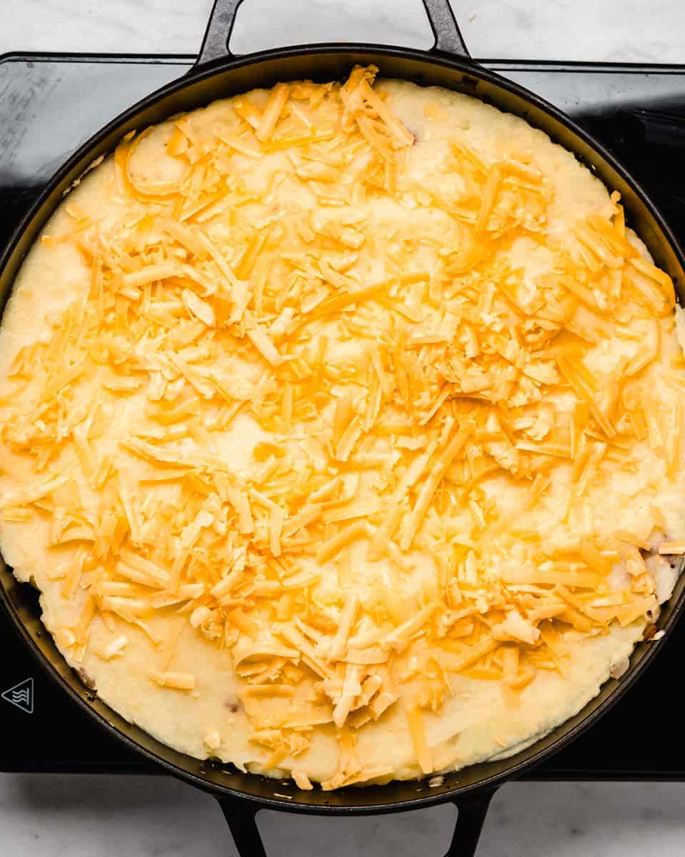 Mashed potatoes and grated cheese on top of the shepherd's pie base in a skillet.