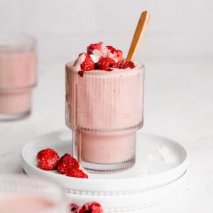 Berry protein smoothie in a glass topped with raspberries and coconut flakes with a spoon tucked inside.