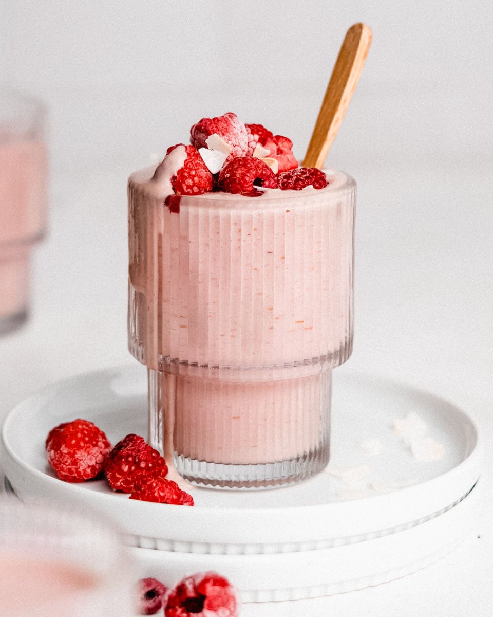 Berry protein smoothie in a glass topped with raspberries and coconut flakes with a spoon tucked inside.