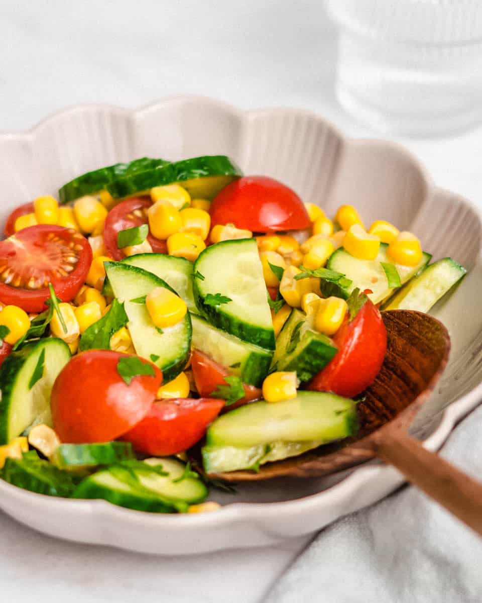 Corn cucumber tomato salad in a bowl and a wooden spoon in it.