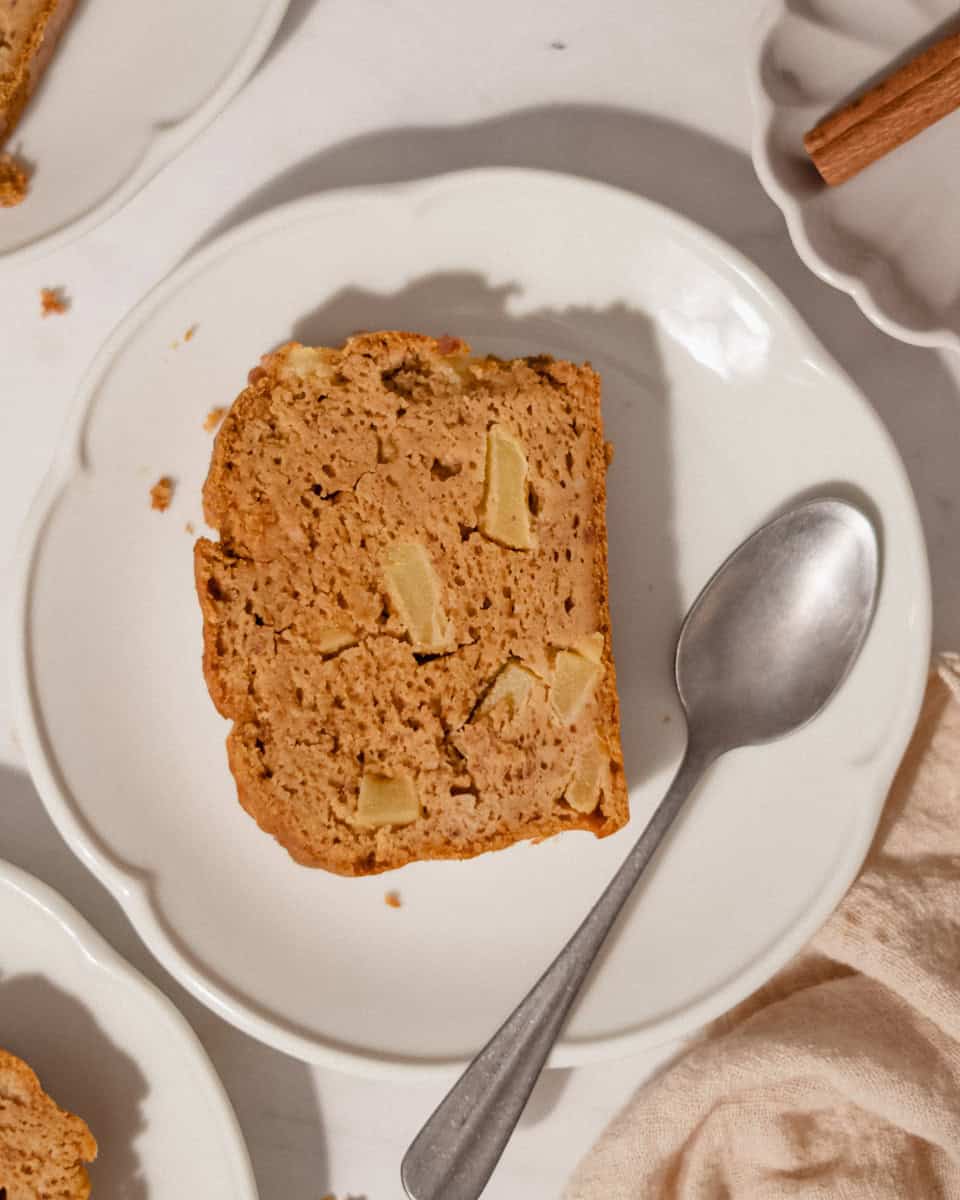 Slice of pumpkin apple bread with apple chunks on a white scalloped plate with a spoon, cinnamon stick on the side.