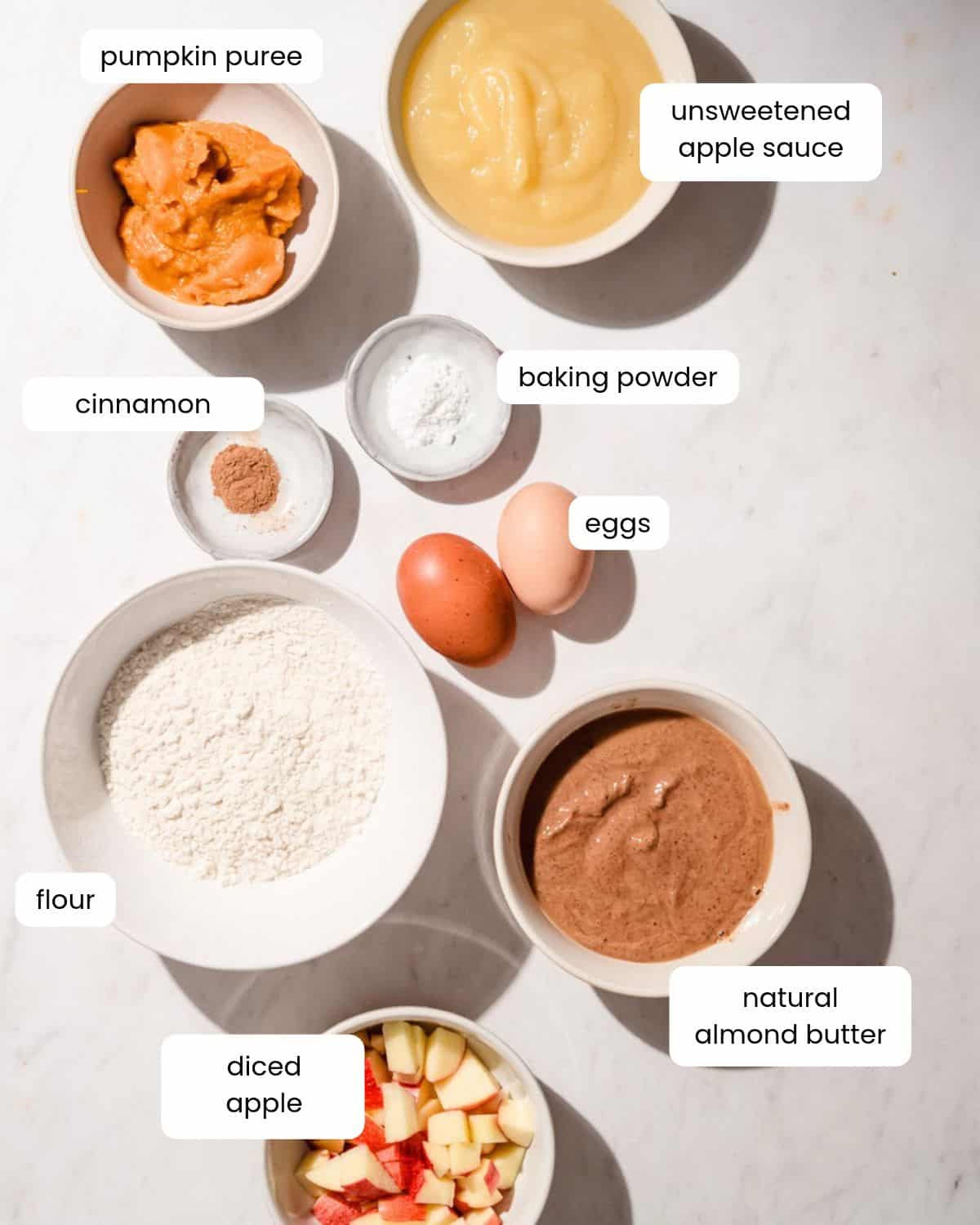 Ingredients laid out in small bowls: pumpkin puree, applesauce, cinnamon, baking powder, two eggs, flour, almond butter and diced apples.