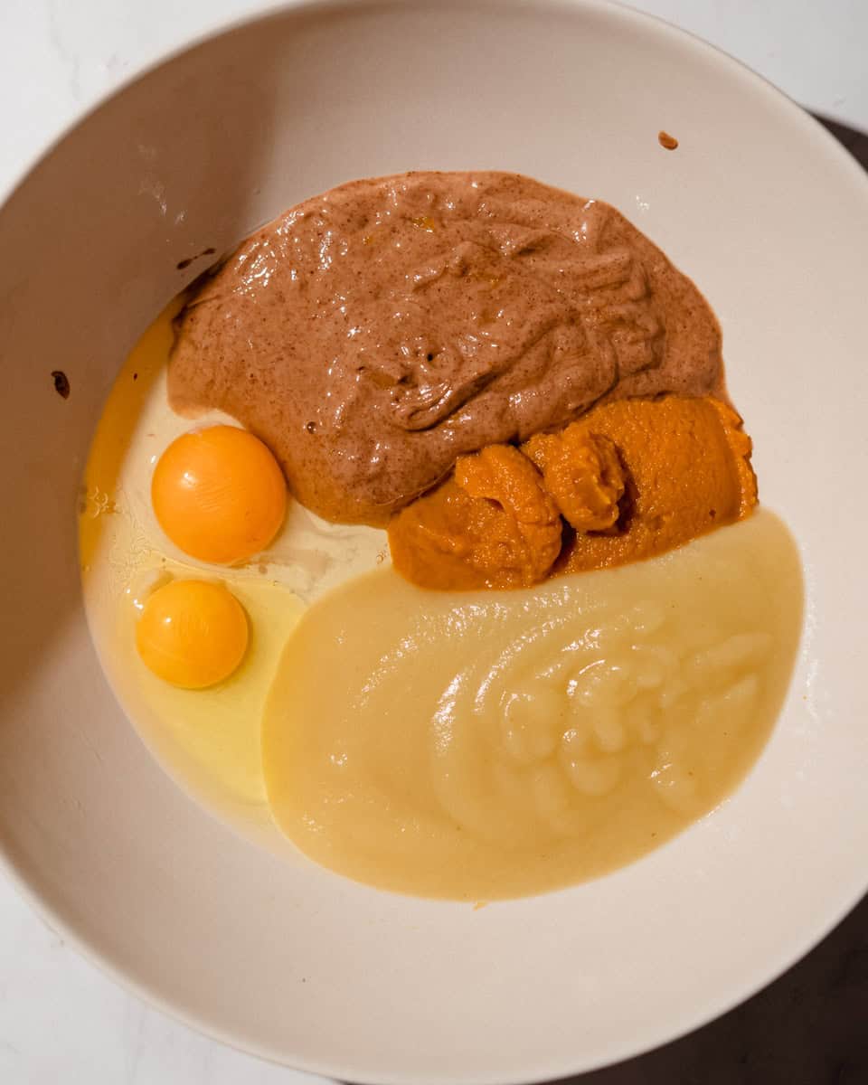 Applesauce, two eggs, pumpkin puree and almond butter added to a large white mixing bowl.