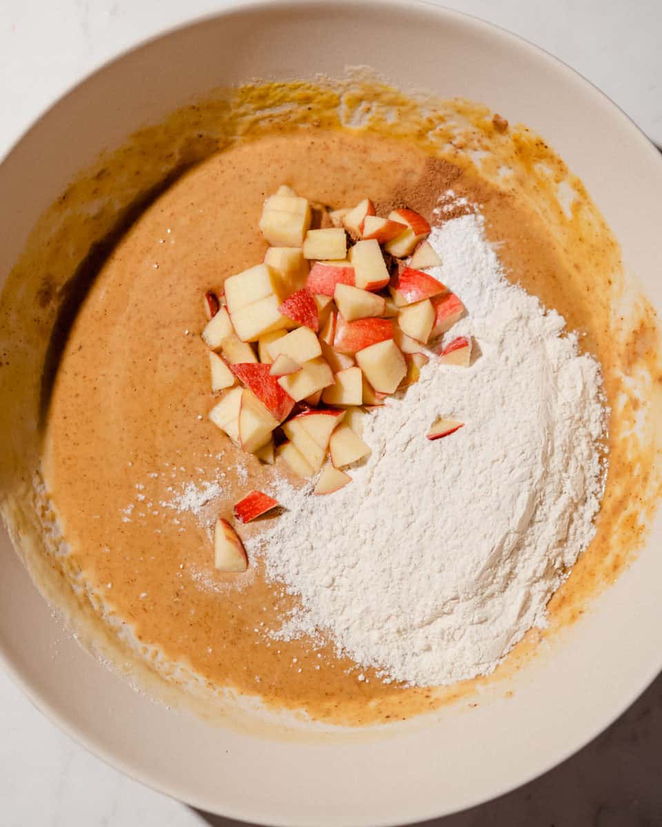 Chopped red apples, flour and cinnamon and baking powder added on top of the wet batter in a mixing bowl.