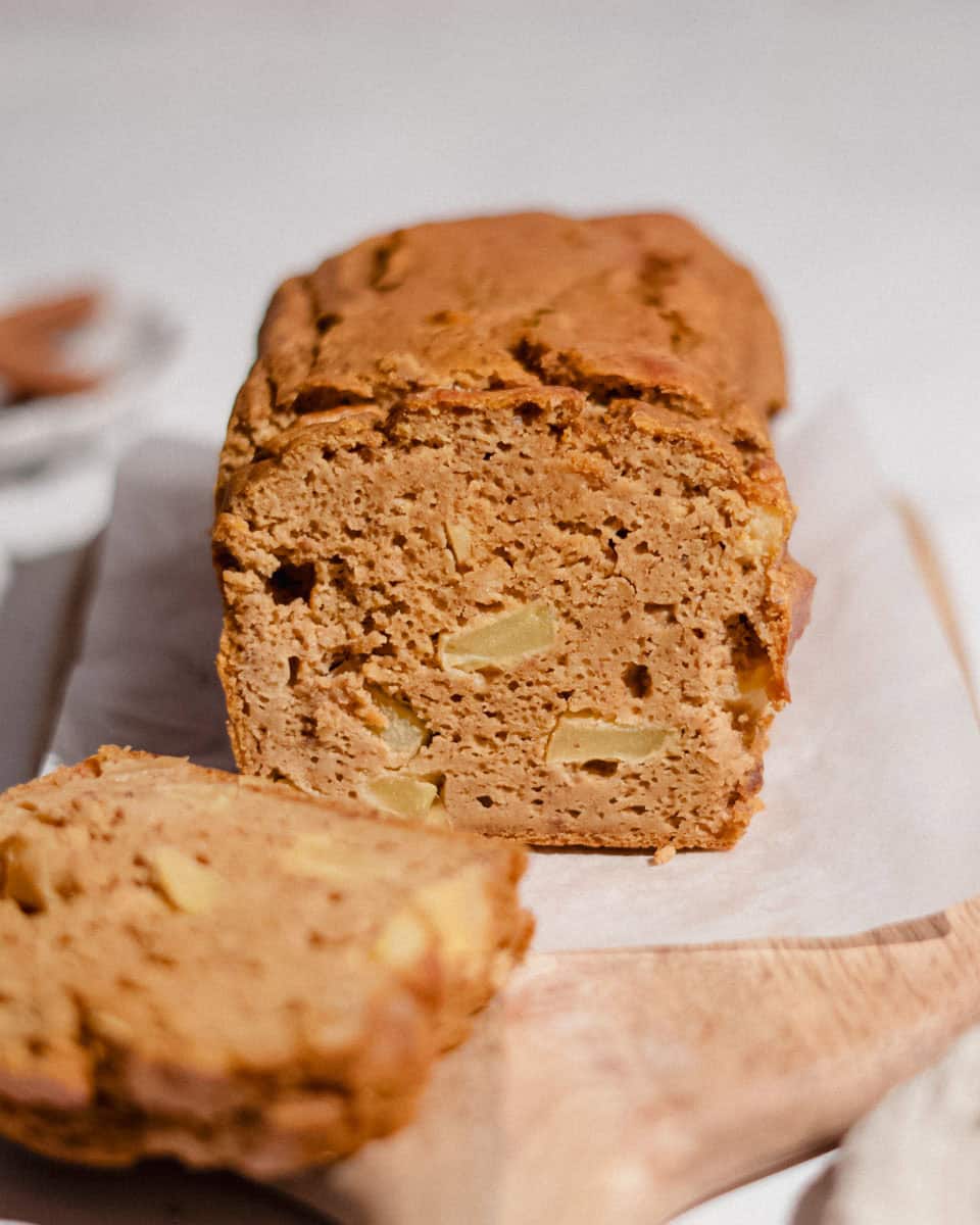 Sliced pumpkin apple bread loaf on parchment paper and a wooden board, showing soft crumb with chunks of apple, one slice in front.