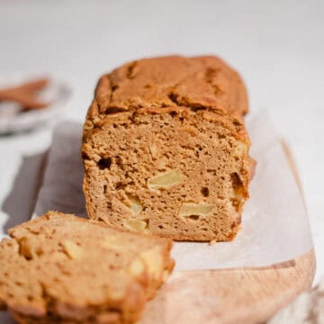 Sliced pumpkin apple bread loaf on parchment paper and a wooden board, showing soft crumb with chunks of apple, one slice in front.