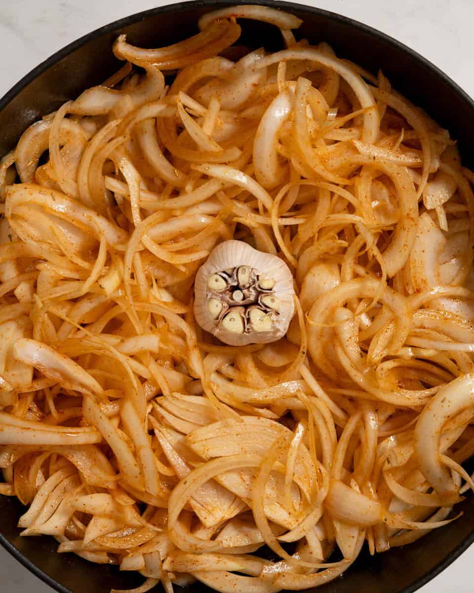 Thinly sliced, raw onions in a dutch oven, a whole garlic bulb placed in the middle.
